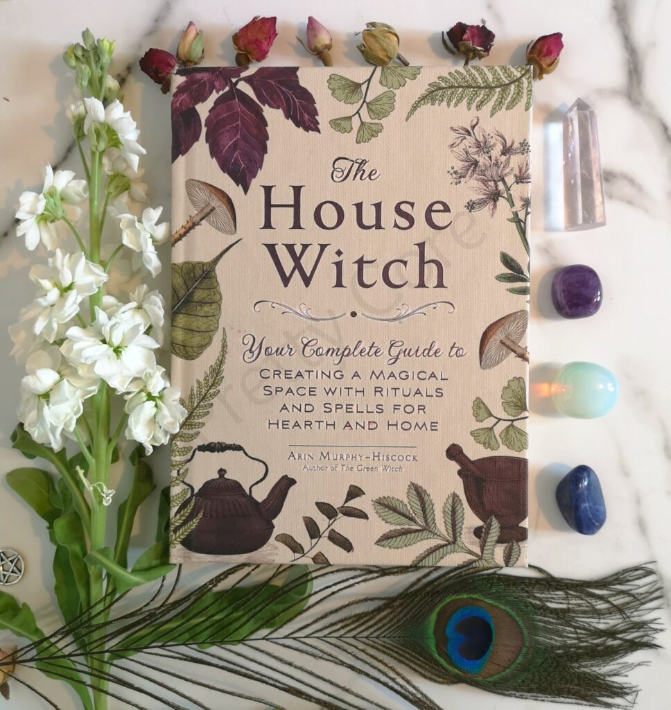 The House Witch Book by Arin Murphy-Hiscock surrounded by slwoers, crystals and a peacock feather.