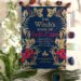 The Witches Book Of Self Care by Arin Murphy-Hiscock in blue and pink, with flowers, peacock feather and crystals surrounding the book