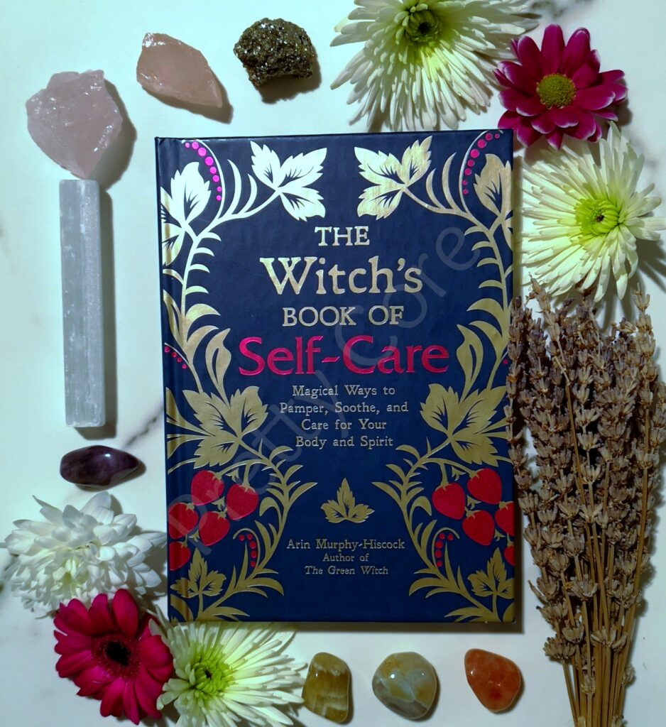 The Witches Book Of Self Care by Arin Murphy-Hiscock in blue and pink, with flowers, lavender and crystals around the book